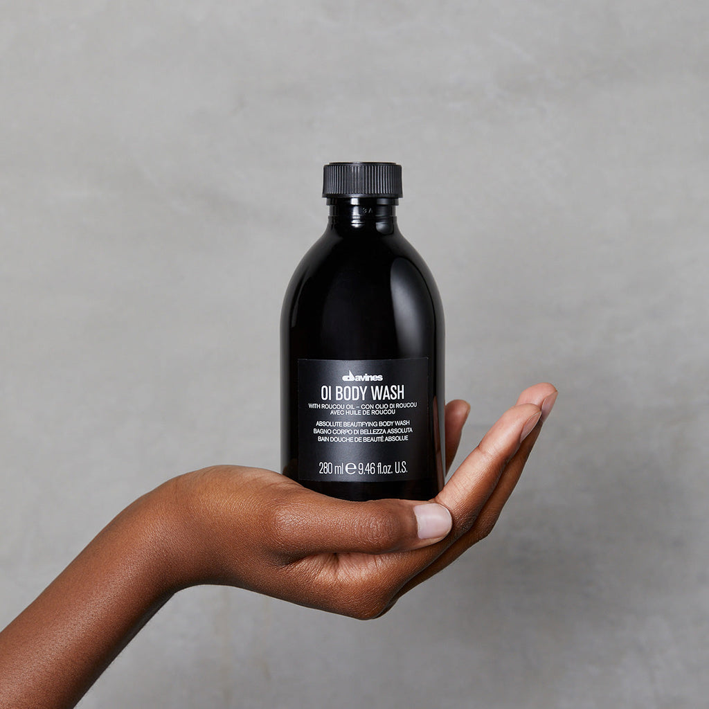 Hand håller en svart flaska med OI/Body Wash mot en grå bakgrund, en miljövänlig kroppstvål från Davines i kompakt design.