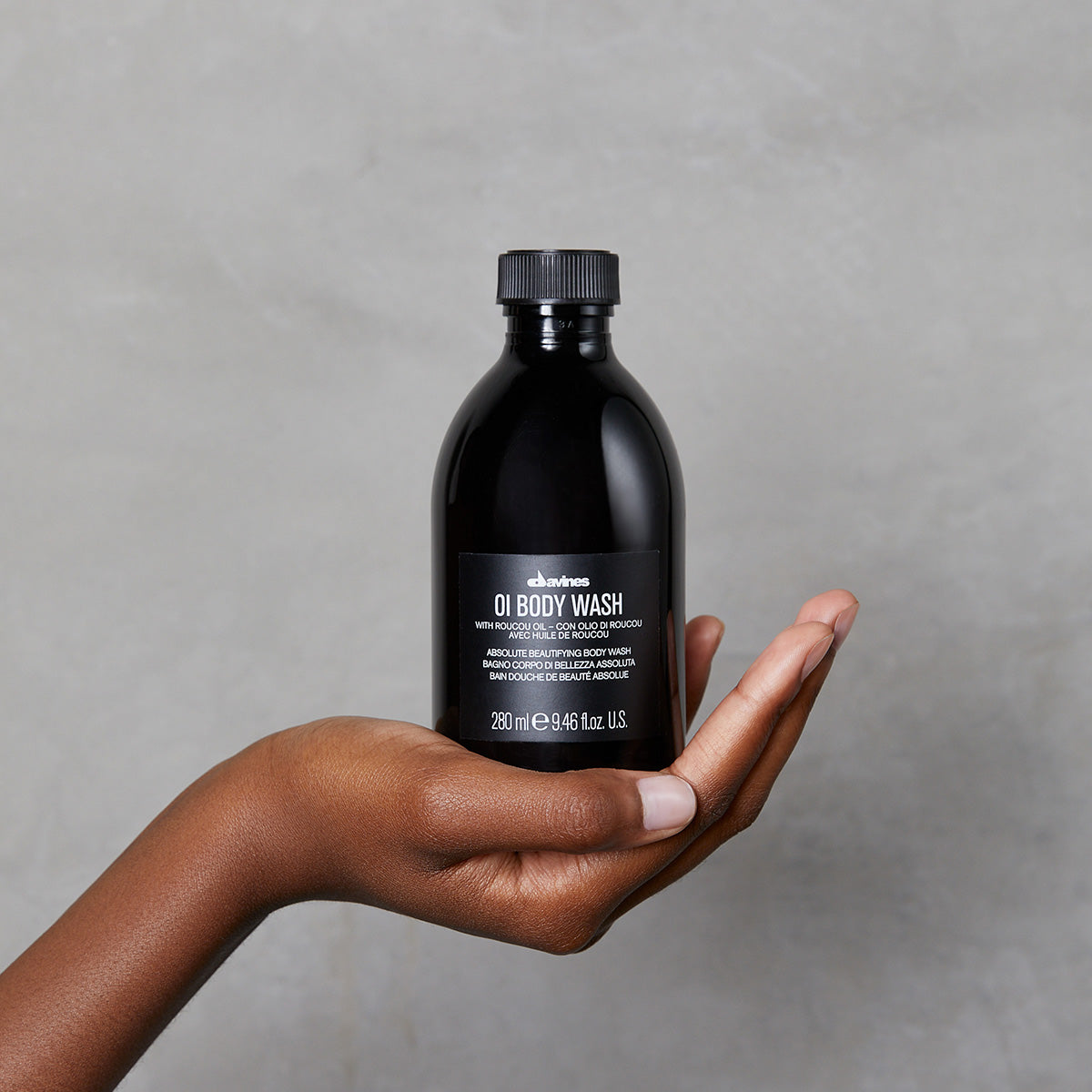 Hand håller en svart flaska med OI/Body Wash mot en grå bakgrund, en miljövänlig kroppstvål från Davines i kompakt design.