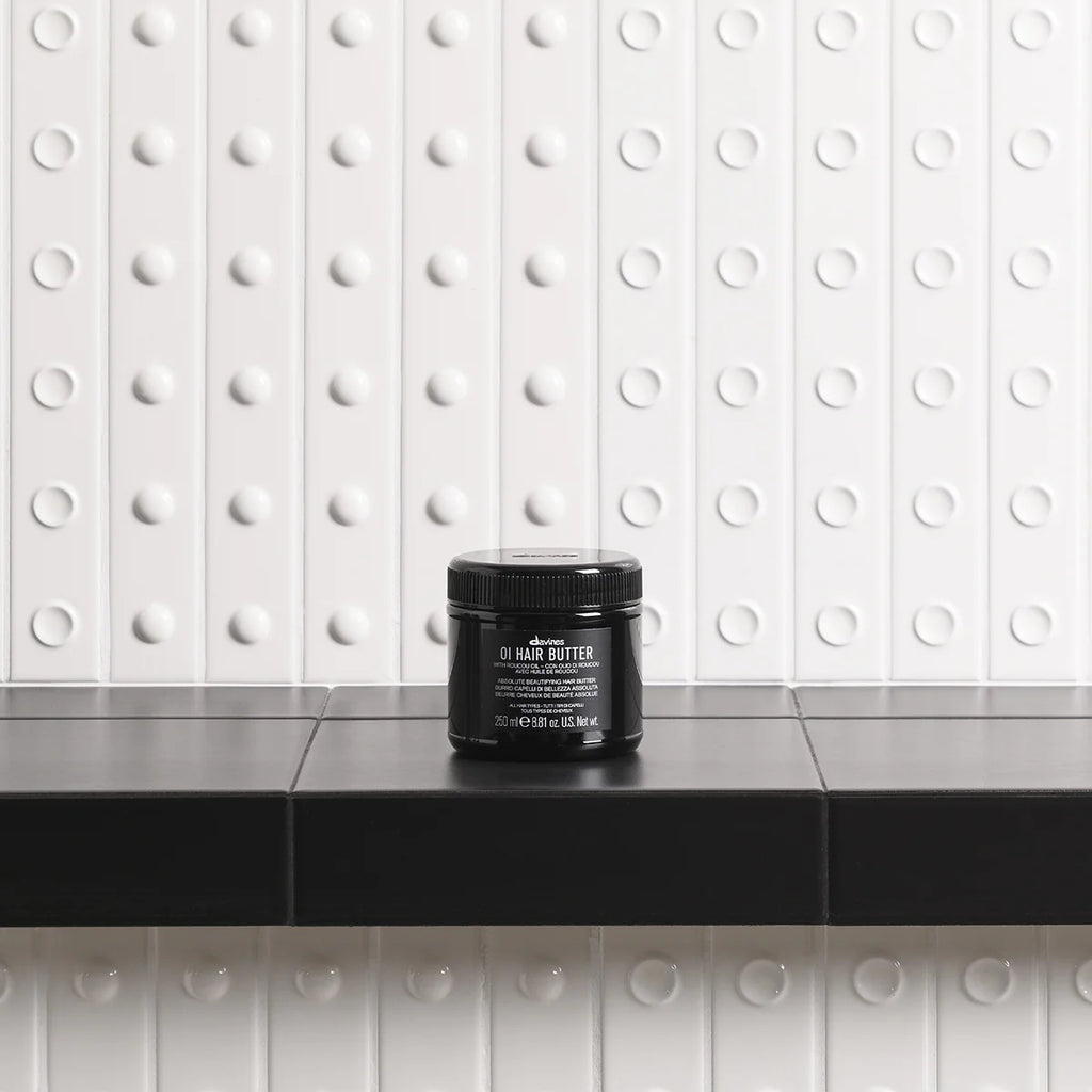 Svart burk med OI/Hair Butter placerad på svart marmorbänk framför vit vägg med prickmönster, stylingprodukt för hårvård.