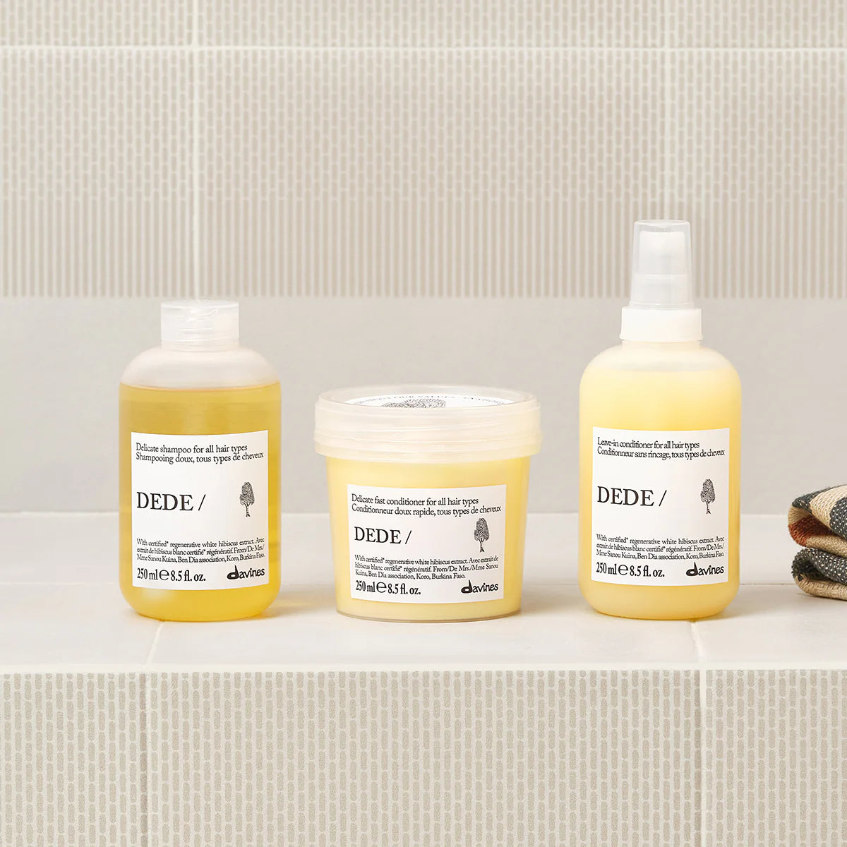 Produkter från Dede Delicate Shampoo-serien med schampo, balsam och hårinpackning i miljövänliga förpackningar på ett badrumshylla i mjuka gula toner.