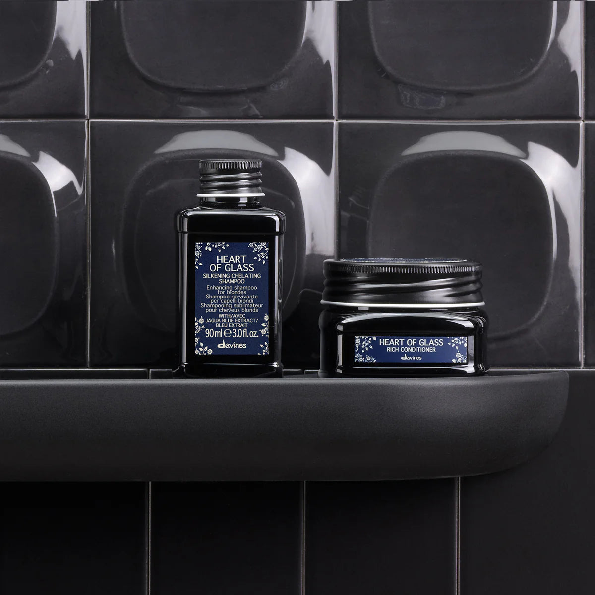 Flaska och burk med Heart of Glass Silkening Shampoo och balsam i mörka behållare på hylla framför svart kakel i badrumsmiljö.