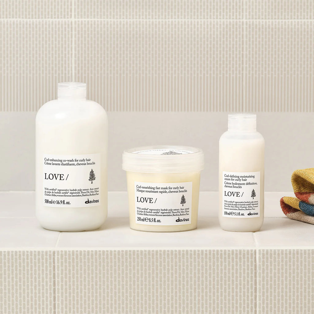 Love Curl Defining Moisturizing Cream i en vit flaska tillsammans med matchande hårvårdsprodukter på en ljus bakgrund, för definiering och vård av lockigt hår.