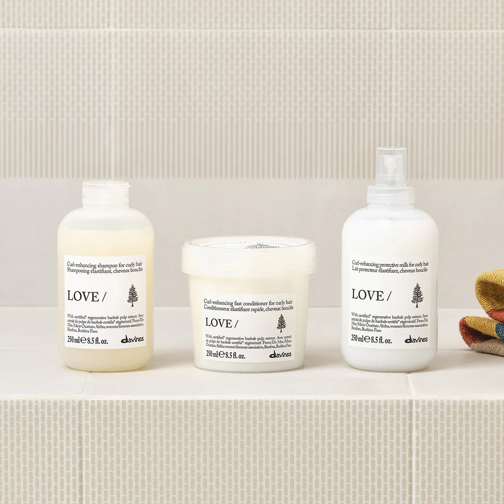 Flera hårvårdsprodukter, inklusive Love Curl Enhancing Protective Milk, placerade på en beige hylla med en enkel bakomliggande vägg, idealiska för styling och vård av lockigt hår.