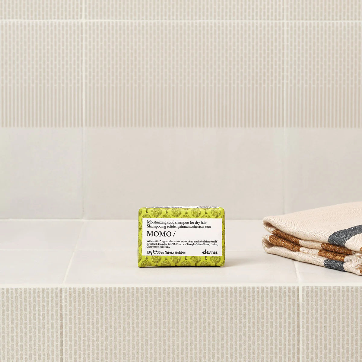 Momo Moisturizing Solid Shampoo Bar för torrt hår, placerad på ett badkarskant intill vikta handdukar, miljövänlig och skonsam schampobar.
