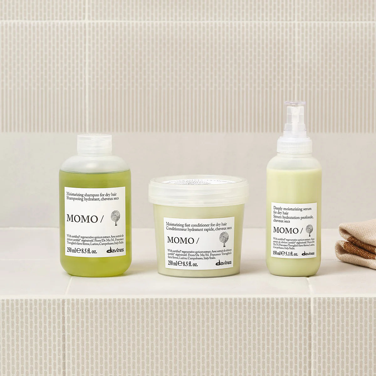 Flaskor med hårvårdsprodukter inklusive Momo Moisturizing Shampoo för torrt hår, placerade på ljus marmorhylla med neutral bakgrund och handduk.