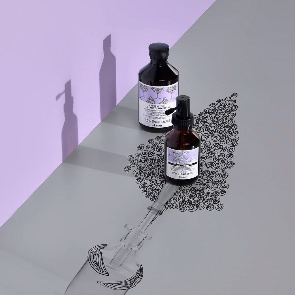 Två flaskor Naturaltech Calming Superactive med lila etiketter står på en yta med en tecknad skiss av hårborste och lockar, en lugnande hårvårdsprodukt från Davines.