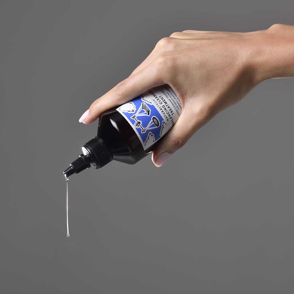 Hand som håller flaska och droppar flytande produkt från Naturaltech Rebalancing Cleansing Treatment mot en grå bakgrund, med fokus på texturen i vätskan.