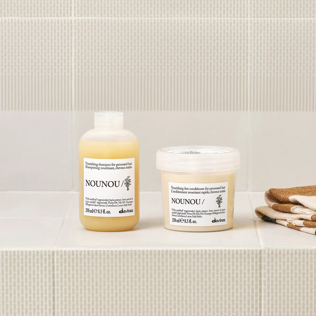 Nounou Nourishing Fast Conditioner i en burk bredvid matchande schampo på ett badrumshylla med beige kakel och vikta handdukar i bakgrunden.