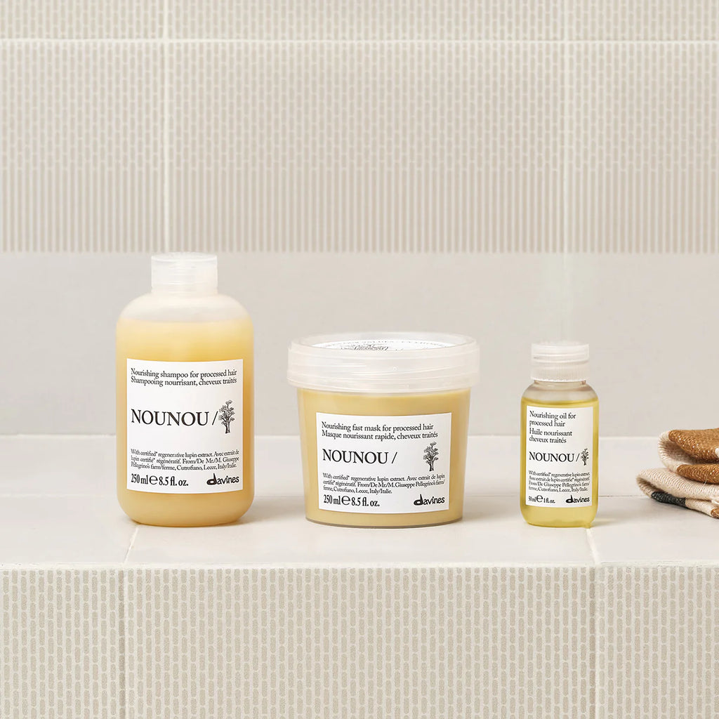 Nounou Nourishing Shampoo i flaska tillsammans med hårinpackning och olja för närande och återuppbyggande behandling av hår, på badrumshylla med beige kakel.