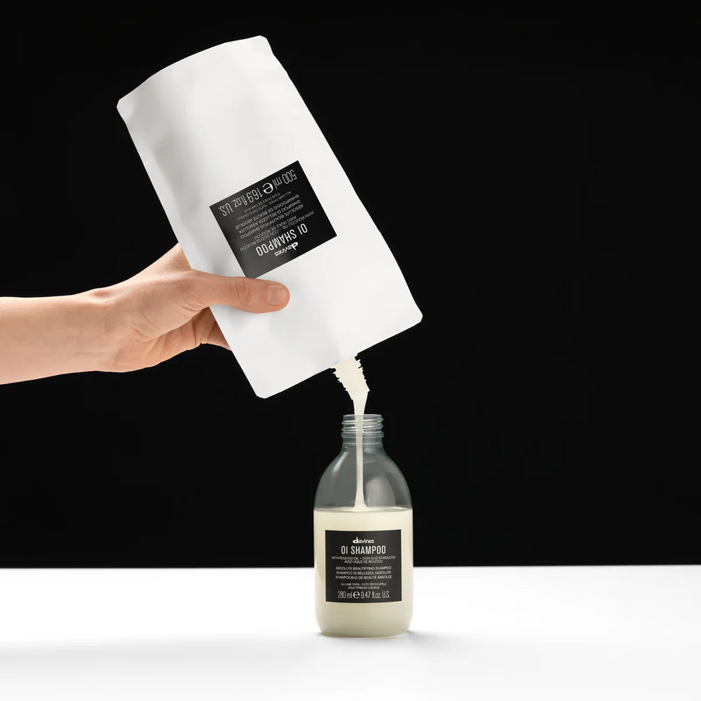Hand häller en refillförpackning med OI/Shampoo schampo i en glasflaska mot svart bakgrund, miljövänlig hårvårdsprodukt.