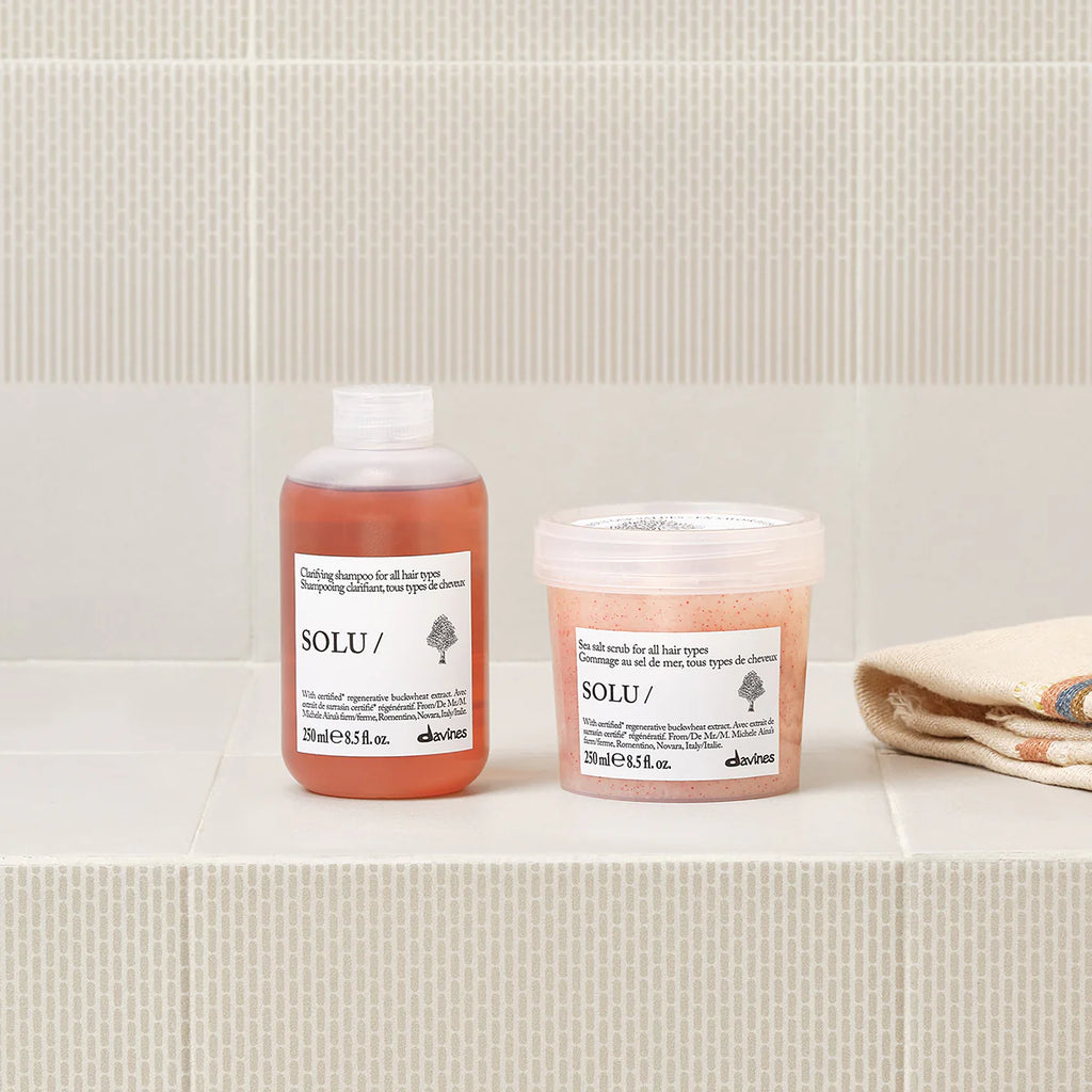 Solu Sea Salt Scrub i en burk tillsammans med en matchande hårprodukt på ett beige klinkergolv i badrumsmiljö, med en vikta handdukar i bakgrunden.