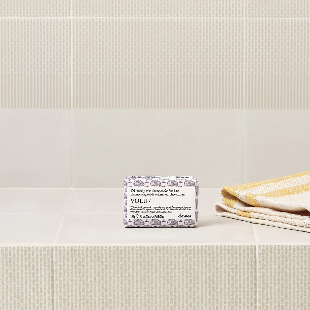 Volu Volumizing Solid Shampoo Bar i miljövänlig förpackning placerad på ett badrumshyllplan med vita och gula handdukar i bakgrunden.