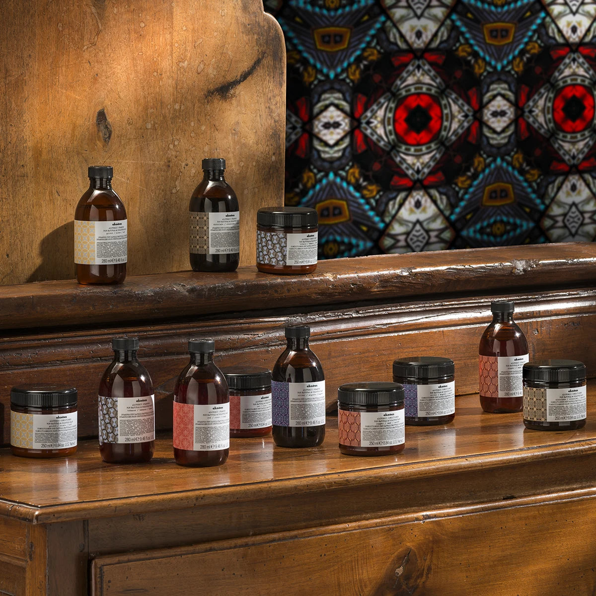 Alchemic Shampoo Silver tillsammans med andra hårvårdsprodukter från Davines i glasflaskor och burkar placerade på ett träbord med färgstarkt bakgrundstyg.