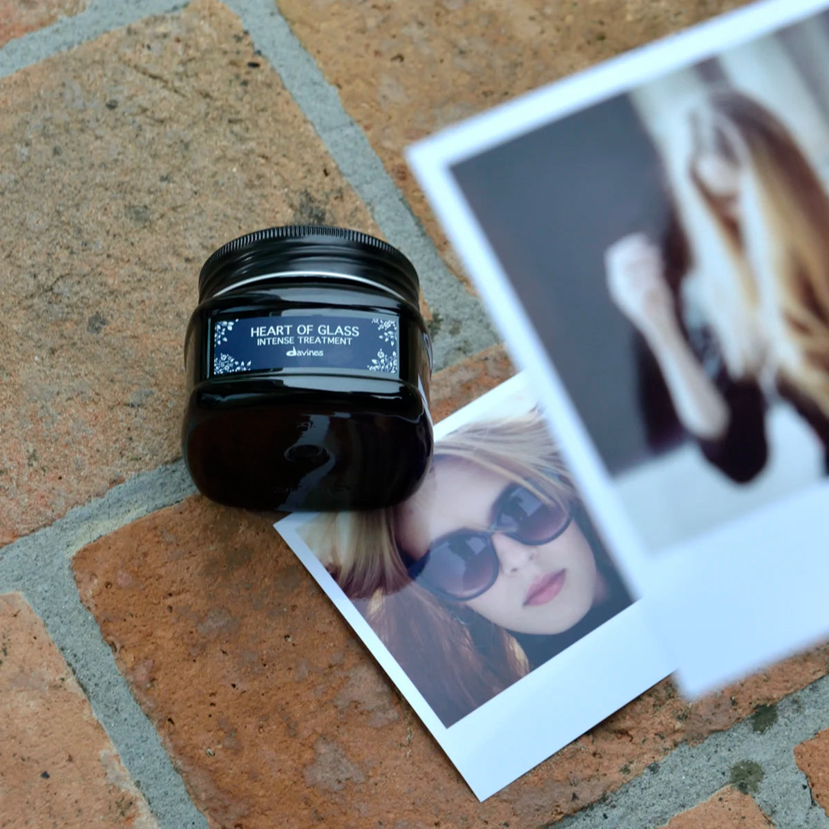 Burk med Heart of Glass Intense Treatment hårinpackning placerad på tegelgolv bredvid två polaroidbilder med porträtt av personer.