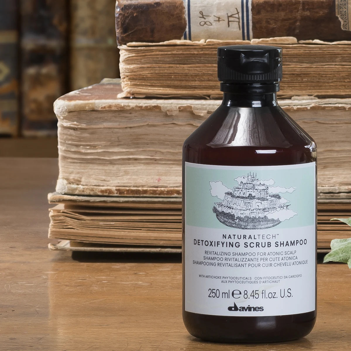 Flaska med Naturaltech Detoxifying Scrub Shampoo stående framför äldre böcker på ett träbord, miljövänligt schampo för hårvård.