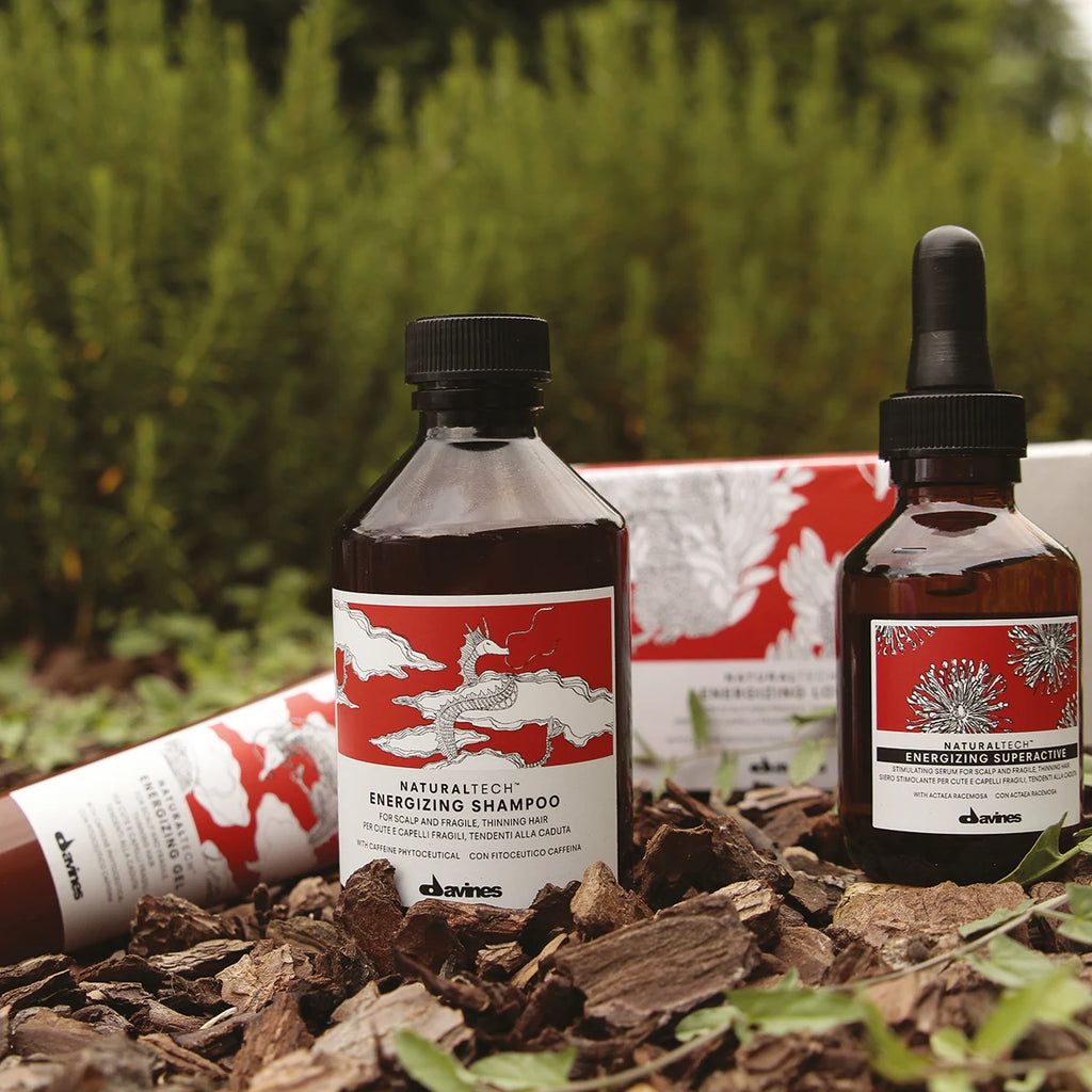Naturlig hårvårdsprodukt i brun flaska med röd och vit etikett, Naturaltech Energizing Gel, stående på marken bland löv och grönska.