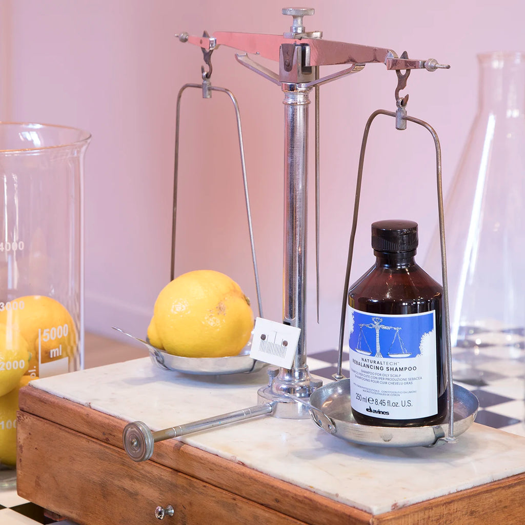 Naturaltech Rebalancing Shampoo i brun flaska balanserad mot en citron på en klassisk metallvåg med glasbägare i bakgrunden, miljövänliga hårvårdsprodukter.