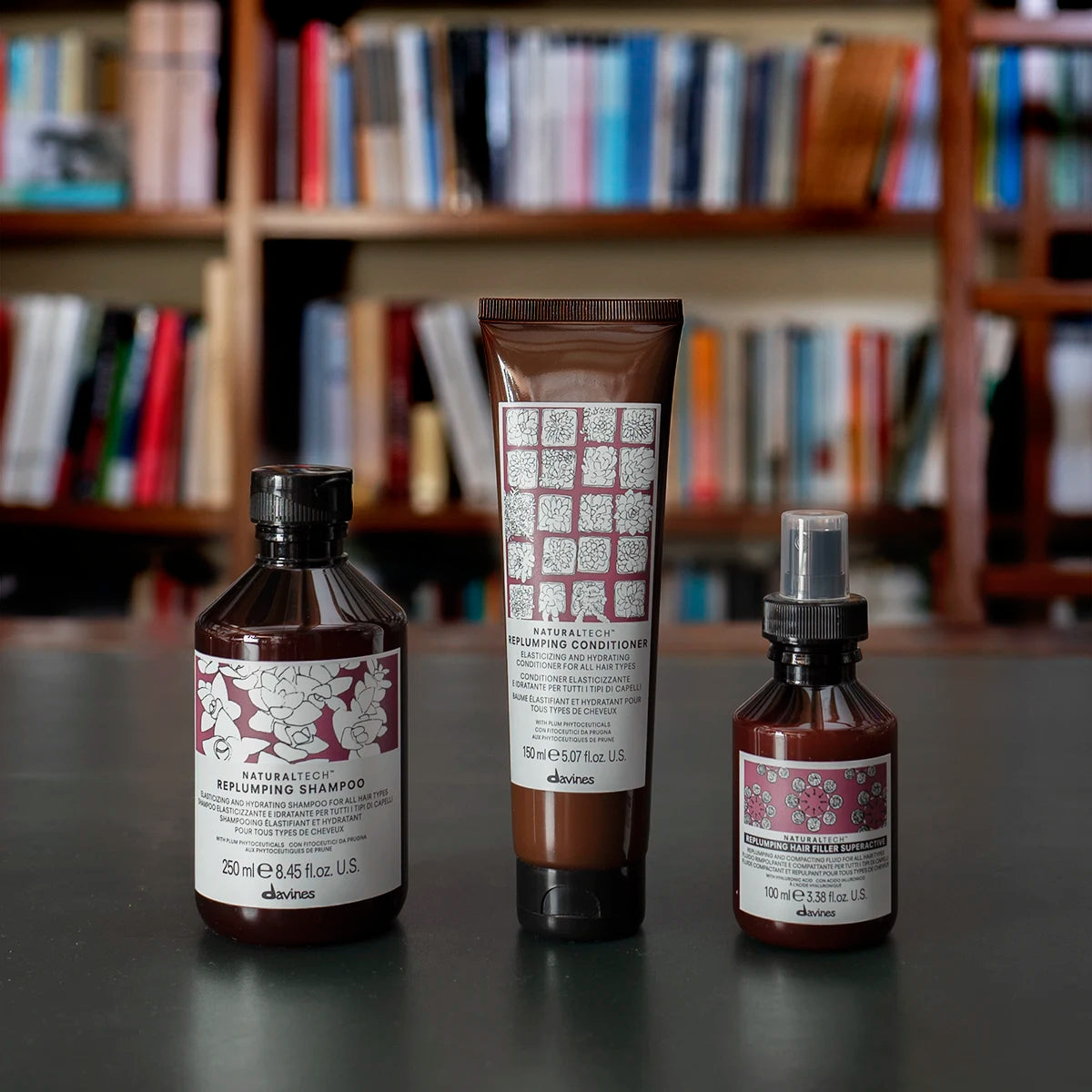 Tre hårvårdsprodukter från Davines med Naturaltech Replumping Hair Filler, inklusive schampo, balsam och en stylingolja, på ett bord framför en bokhylla.