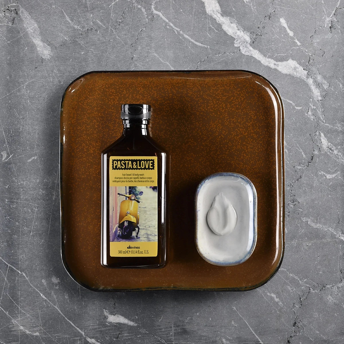 Flaska med Pasta & Love Hair Beard & Body Wash intill ett brunt fat med kräm på en tvål, placerade på en grå marmoryta.