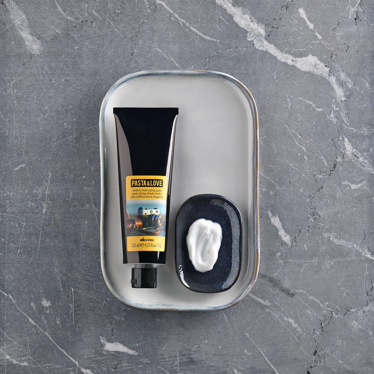 Pasta & Love Medium Hold Styling Paste i svart tub bredvid en svart skål med krämig, vit stylingprodukt på en metallbricka mot en marmorytpa.