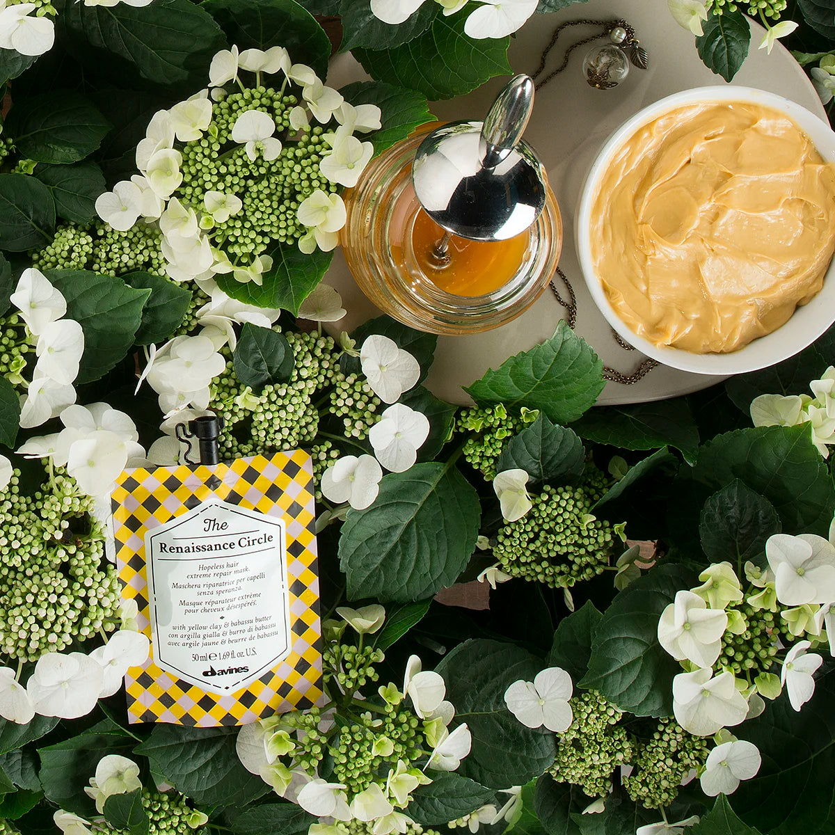 The Renaissance Circle hårinpackning i en gul och svart förpackning omgiven av vita blommor och en skål med krämig hårvårdsprodukt på ett grönt bladunderlag.