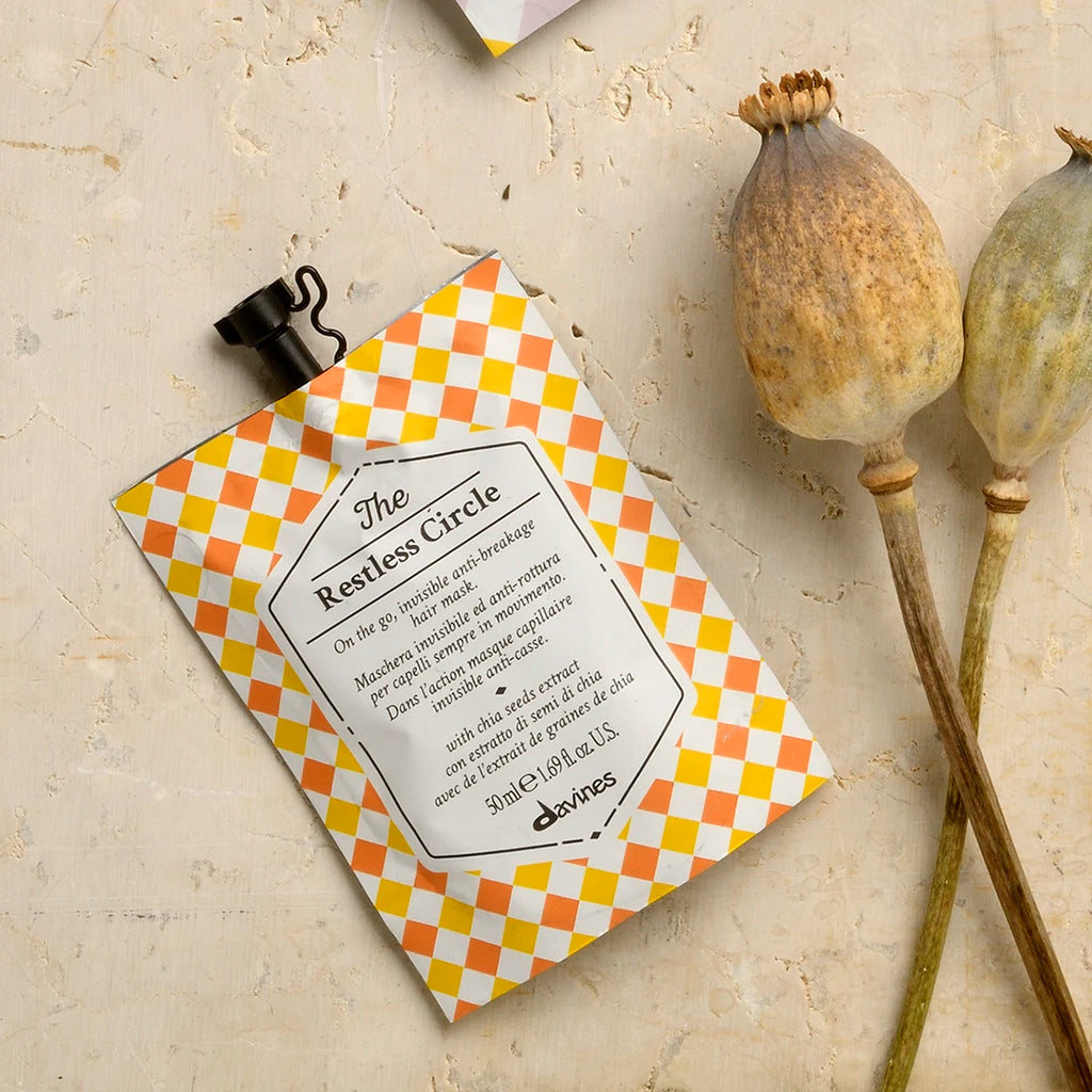 Förpackning med miljövänliga hårvårdsprodukten The Restless Circle från Davines, en löddrande hårinpackning i färgglad design liggande bredvid torkade vallmokapslar.