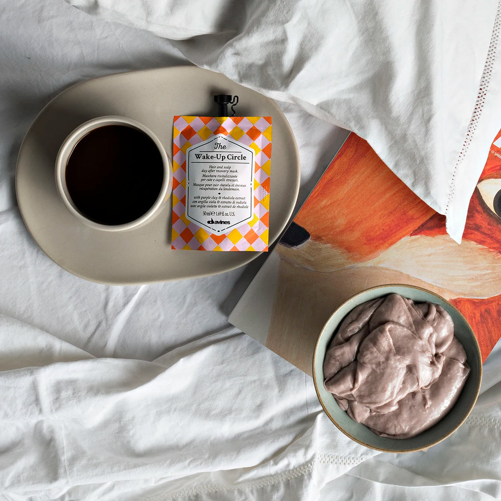 The Wake-Up Circle hårinpackning från Davines placerad på en tallrik bredvid en kopp kaffe och en skål med krämig mousse på en vit bäddad säng med en bok under.